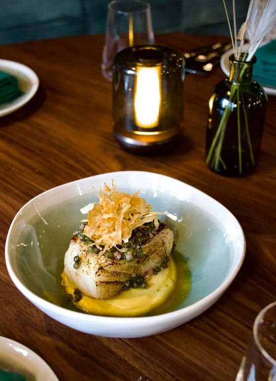 A beautifully plated piece of fish topped with crispy garnish, served on a creamy sauce in a light blue bowl, surrounded by elegant dining decor.