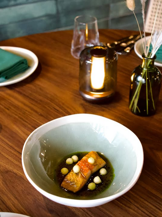 A beautifully plated dish featuring a piece of fish on a bed of green sauce, garnished with small white spheres, served in a pale green bowl on a wooden table.