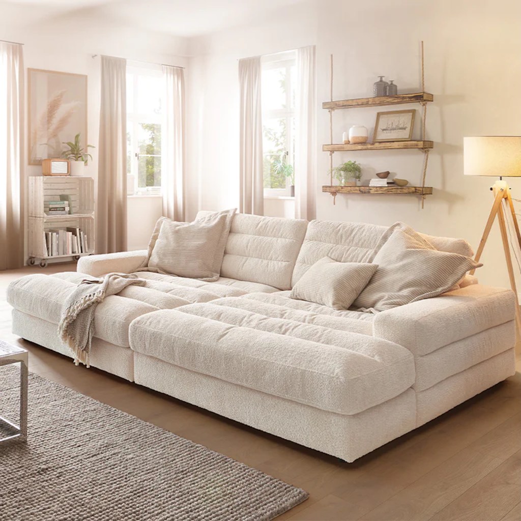 Modern living room featuring a plush, light-colored sectional sofa with decorative pillows, a woven throw blanket, a decorative rug, and natural light coming through large windows.