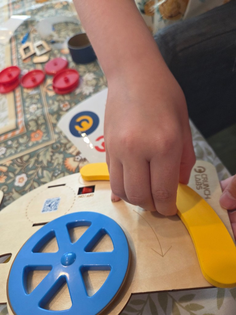 A child's hand manipulating a yellow lever on a wooden activity board with a blue wheel, surrounded by various colorful game pieces.