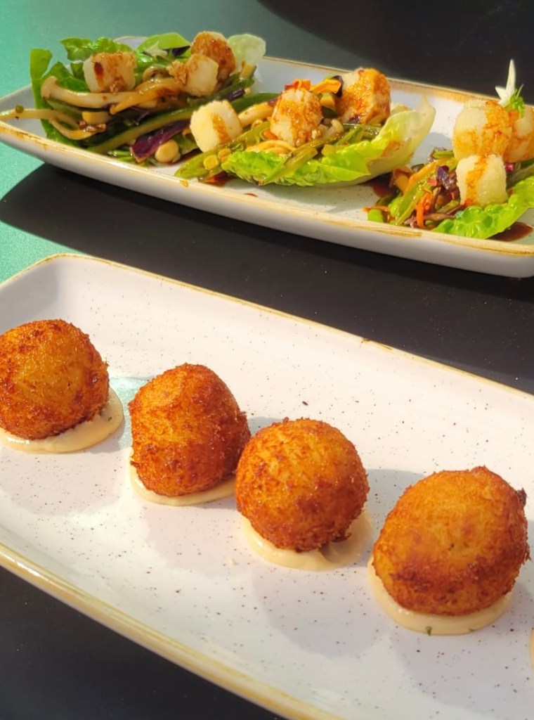 A plate with four golden brown croquettes accompanied by a creamy sauce, placed in front of a colorful salad topped with grilled shrimp and various vegetables.