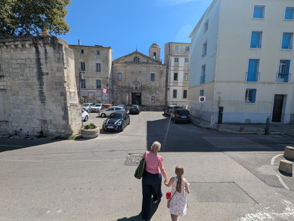 A woman and a girl walking hand in hand on a sunny street, with historic buildings and parked cars in the background.