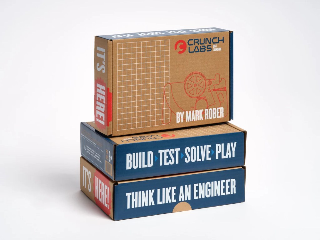 Stack of three educational boxes from Crunch Labs, designed by Mark Rober, featuring texts like 'It's Here!', 'Build Test Solve Play', and 'Think Like an Engineer'.