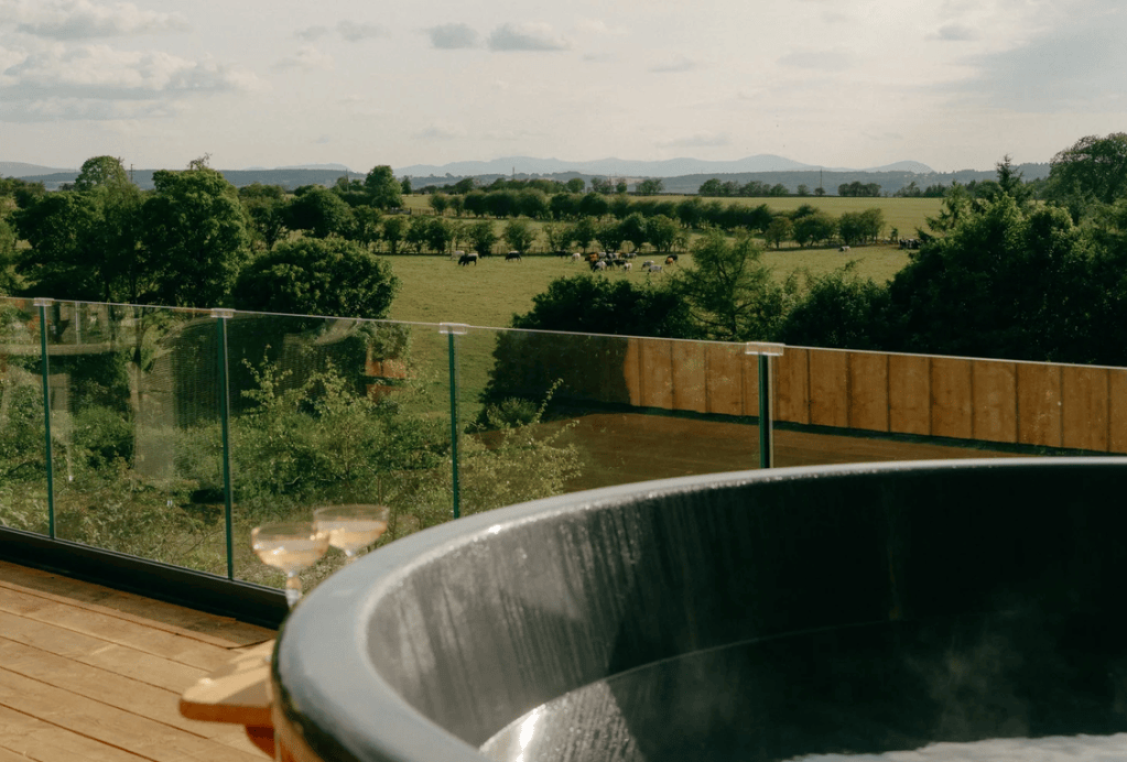 View of a hot tub with champagne glasses, overlooking a scenic landscape of fields and mountains in the distance.