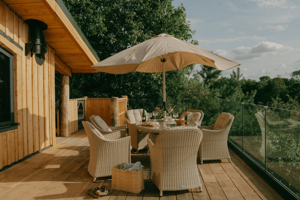 A cozy outdoor dining area featuring a round table with glasses and a bottle, surrounded by wicker chairs and an umbrella, set on a wooden deck with lush greenery in the background.