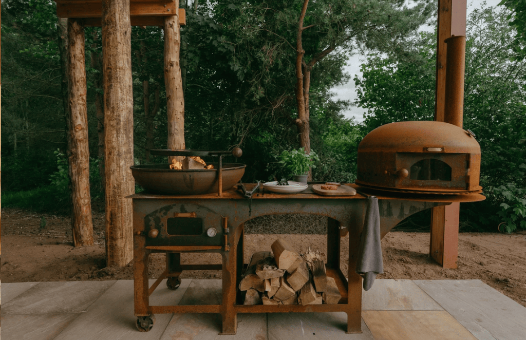 An outdoor cooking station featuring a rusted metal grill with a bowl of flames, a pizza oven, and a stack of firewood, surrounded by trees.