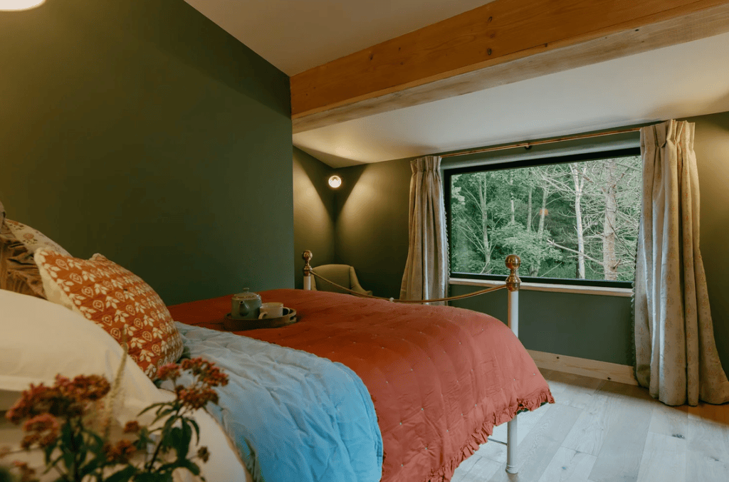 Cozy bedroom featuring a patterned pillow, a red and blue quilt, and a window overlooking a lush green forest.
