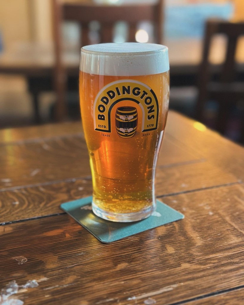 A glass of Boddingtons beer with a creamy white head, placed on a wooden table with a blue coaster.